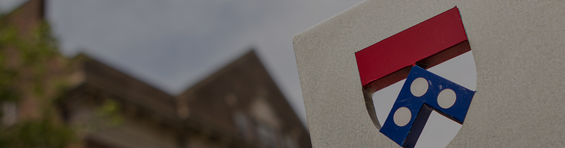 EMBA_Contact_1900x500 A partial view of a university emblem on a sign with a blurred building and tree in the background.