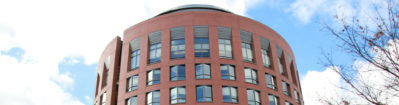 MBA_Visiting_Hero_1900x500-2-1900x500-1900x500 (1) A red brick building with a cylindrical shape, featuring multiple windows in a modern architectural style against a partly cloudy sky.