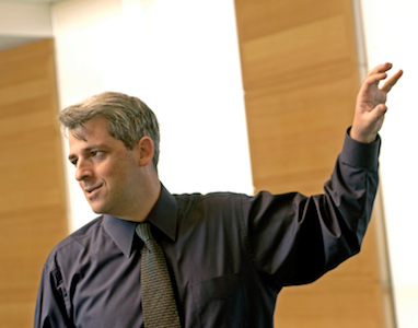 A person in business attire gestures upwards while appearing to give a presentation or lecture, with a neutral background.