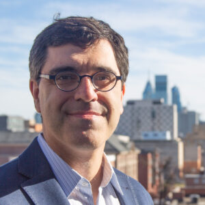 Headshot of a person wearing glasses and a suit, standing outdoors with a cityscape in the background under a blue sky.
