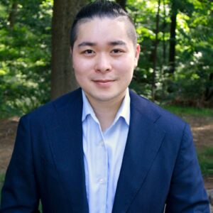 Headshot of a person in a blue suit and light blue shirt standing in a forested area.